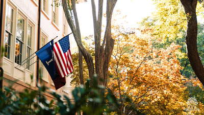 The American and South Carolinian flag on the President’s House on a sunny day with tree and gates mark.