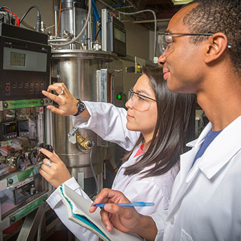 Two student reserrchers in front of machinery taking a reading wearing safety goggles and lab coats.