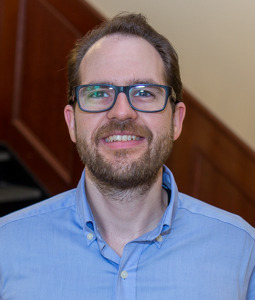 Nicolas wears a blue oxford shirt and smiles at the camera